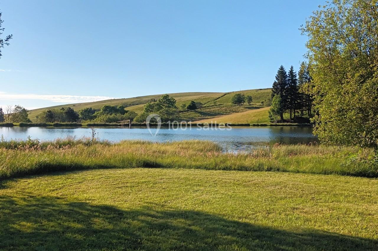 Paysage avec un lac entouré de prairies, d'arbres et de collines sous un ciel bleu dégagé.