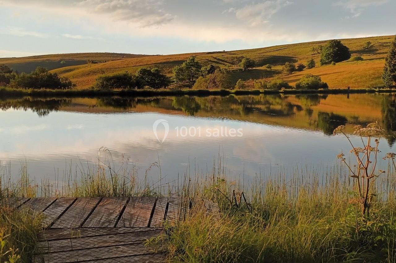 Vue d'un lac calme entouré de collines verdoyantes et de végétation, avec un ponton en bois au premier plan.