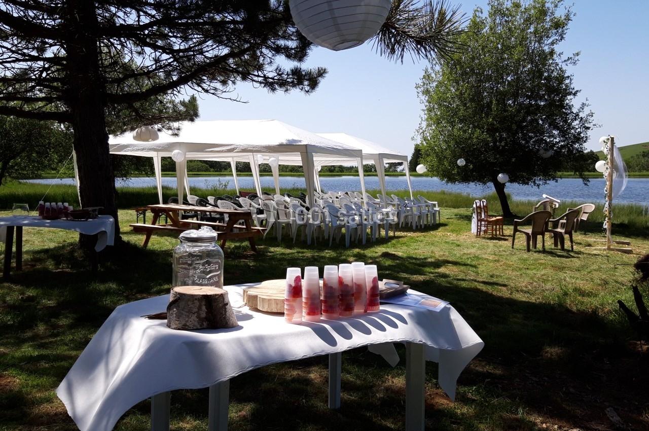 Tables dressées en extérieur avec des boissons, des chaises blanches sous une tente et un lac en arrière-plan.