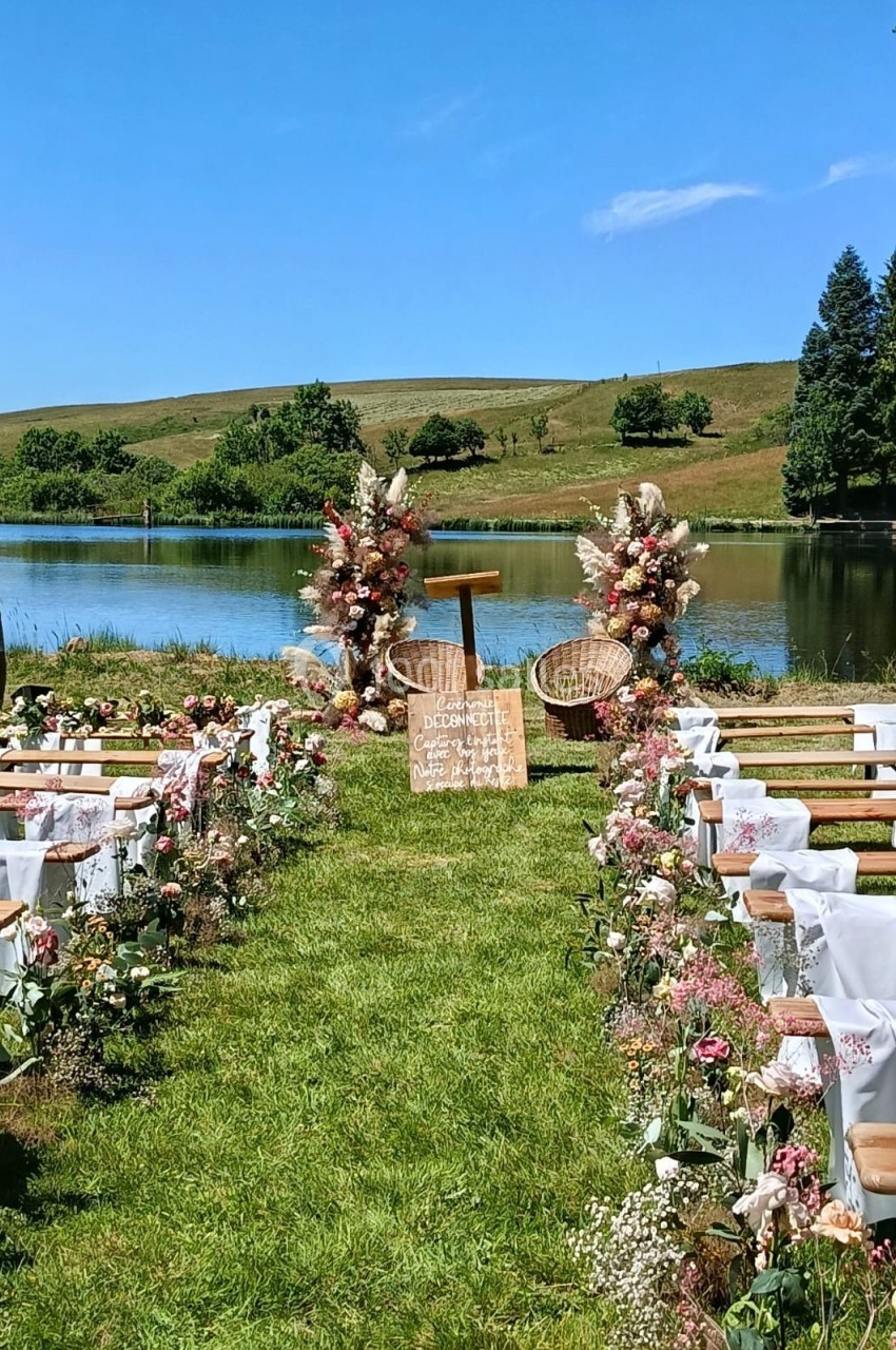 Allée décorée de fleurs menant à un autel en plein air près d'un lac, entouré de collines verdoyantes.