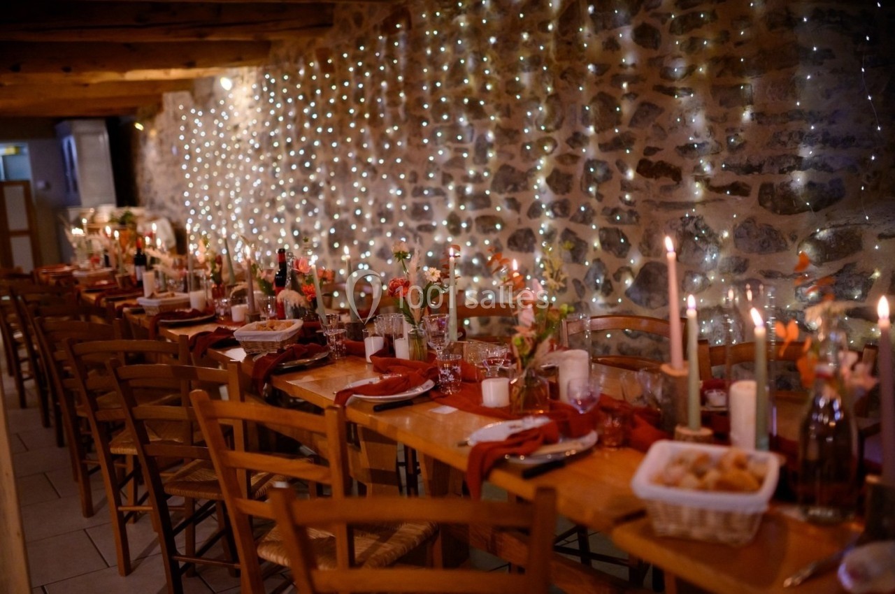 Salle rustique décorée pour un dîner, avec longues tables en bois, bougies allumées et guirlandes lumineuses sur un mur en…