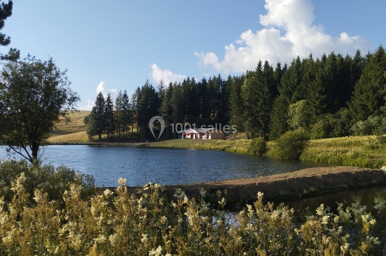 Vue d'un lac entouré de végétation, avec une maison isolée en bordure d'une forêt sous un ciel bleu.
