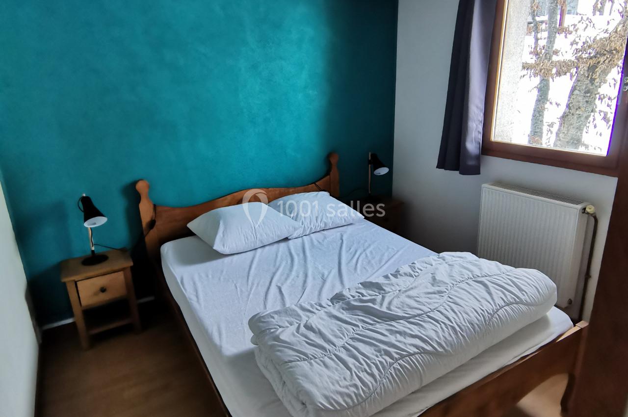 Chambre avec lit double en bois, mur turquoise, poutres apparentes, fenêtre donnant sur un arbre, parquet au sol.