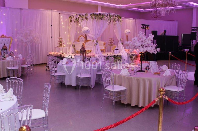 Salle de réception décorée avec des tables rondes, des chaises transparentes et une scène ornée de fleurs et lumières.