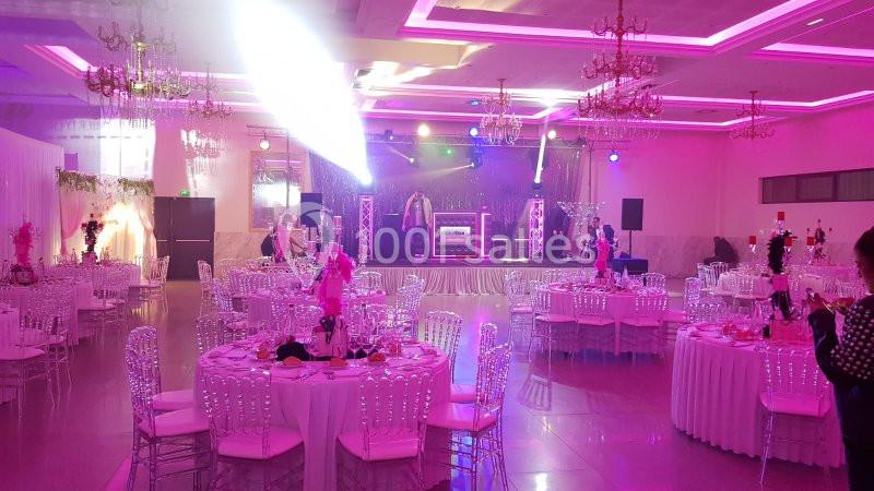 Salle de réception décorée avec des tables rondes, des chaises transparentes et une scène éclairée par des projecteurs.