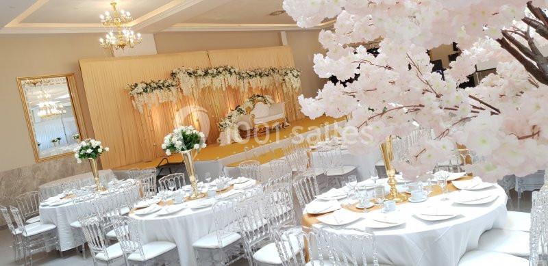 Salle de réception décorée avec des tables rondes, des chaises transparentes et des arrangements floraux blancs et roses.