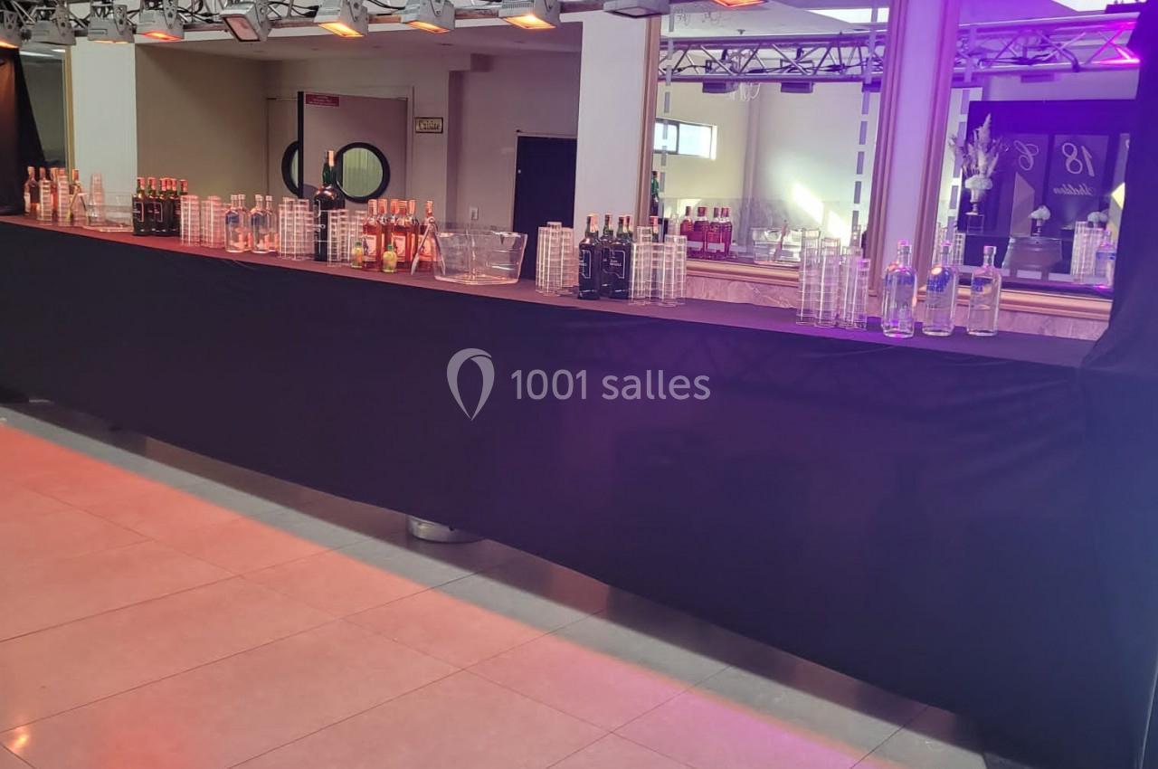 Bar long avec nappes noires, verres, bouteilles et éclairage violet dans une salle lumineuse avec miroirs.