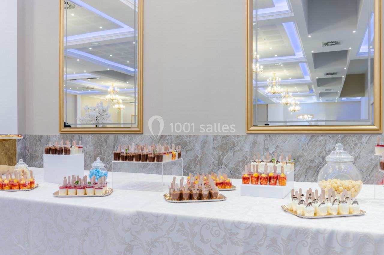Table garnie de desserts variés en verrines et bonbons, disposée dans une salle élégante avec miroirs et lustres.