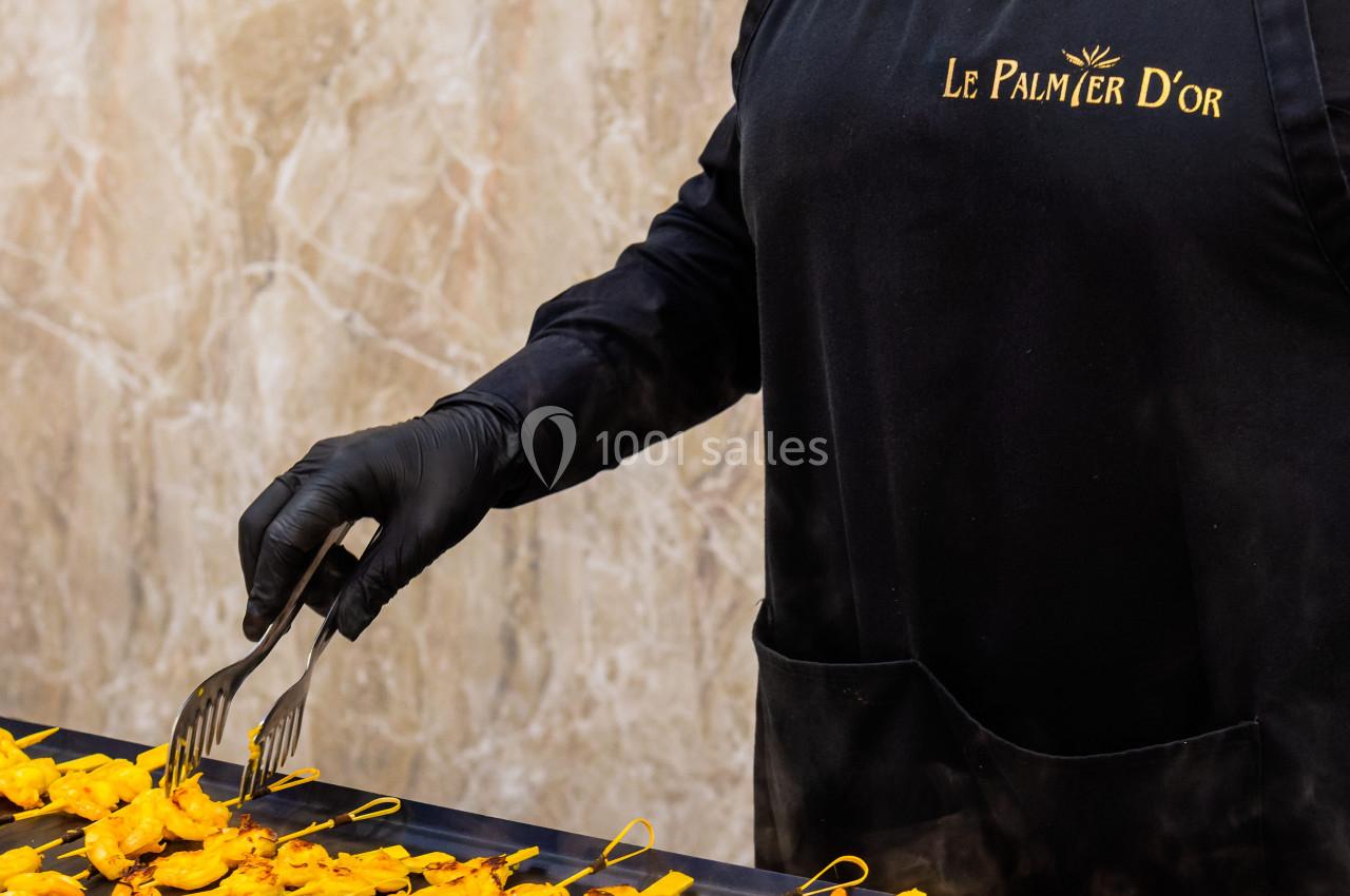 Un serveur en tablier noir dispose des brochettes sur une table en utilisant des pinces, devant un mur en marbre beige.