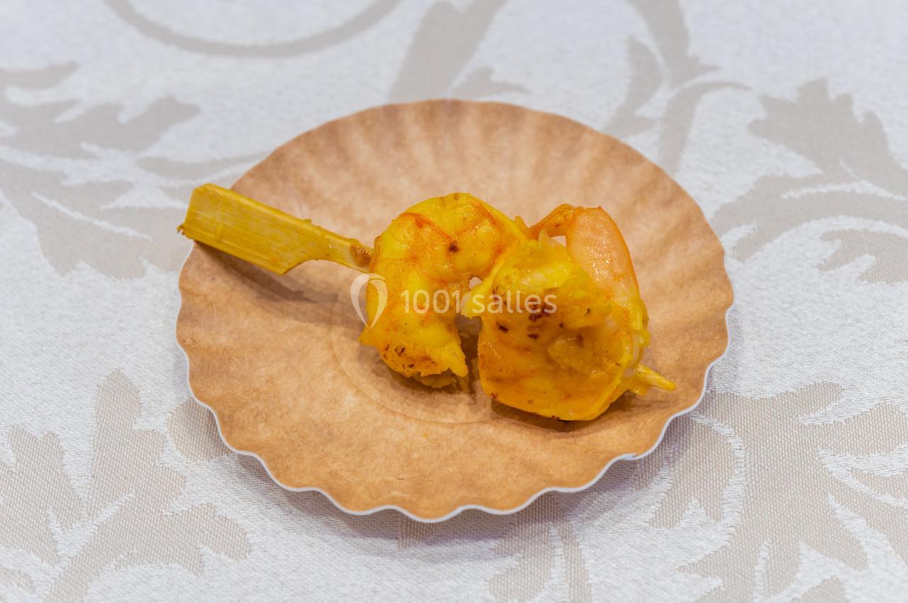 Deux crevettes grillées sur un bâtonnet, présentées sur une assiette en papier beige avec une nappe en arrière-plan.