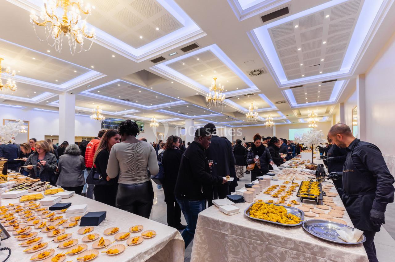 Salle lumineuse avec des convives se servant à un buffet varié, sous des lustres élégants.