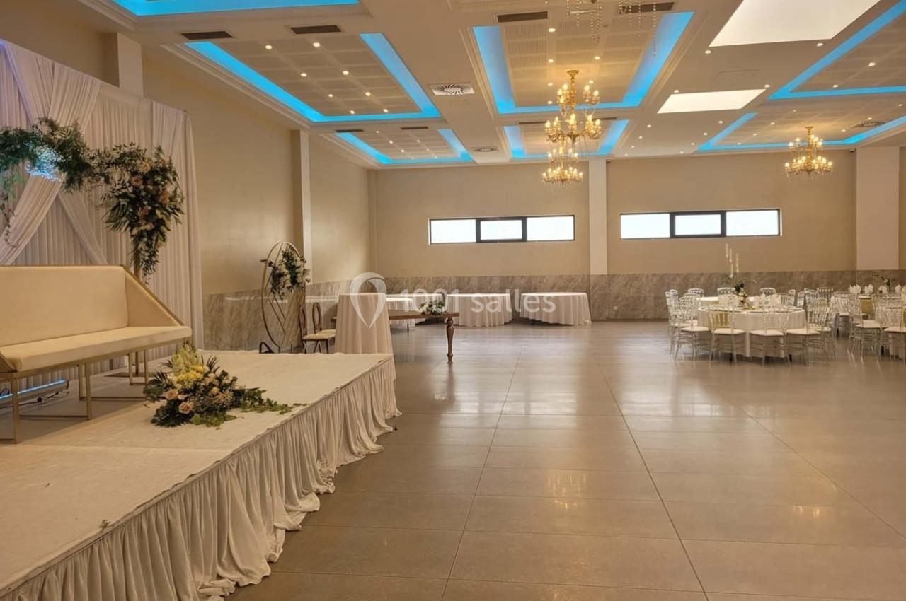 Salle de réception lumineuse avec tables dressées, scène décorée de fleurs et lustres suspendus au plafond.