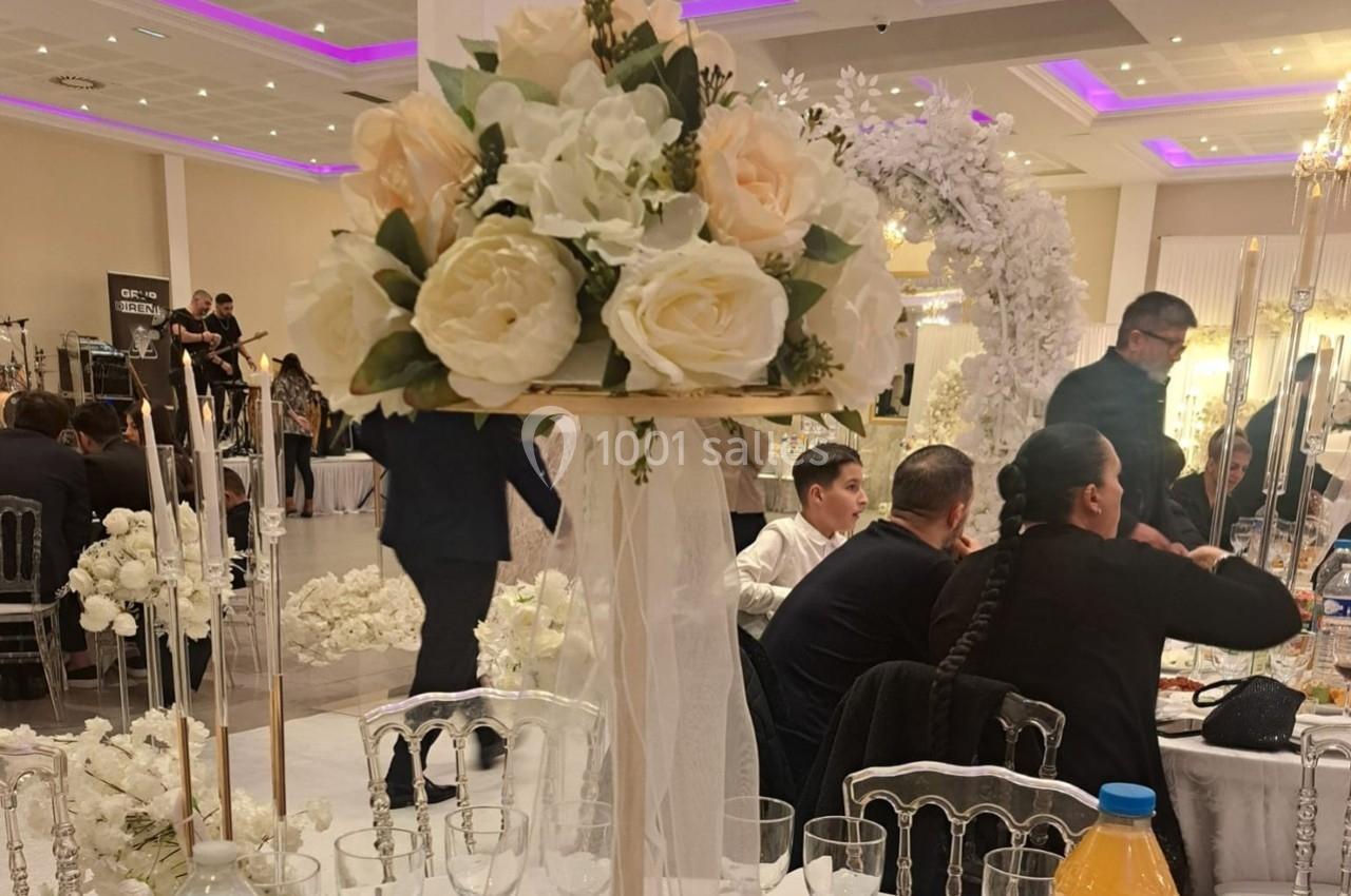 Centre de table floral avec roses blanches et pêche, dans une salle de réception élégante avec invités en arrière-plan.