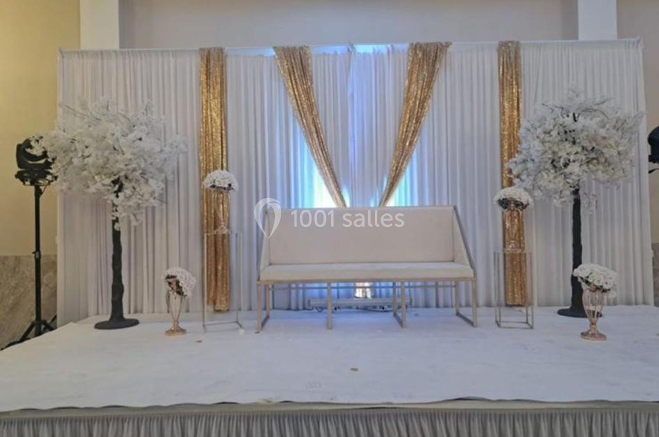 Décor de scène avec rideaux blancs et dorés, banc blanc, arrangements floraux blancs et éclairage doux.