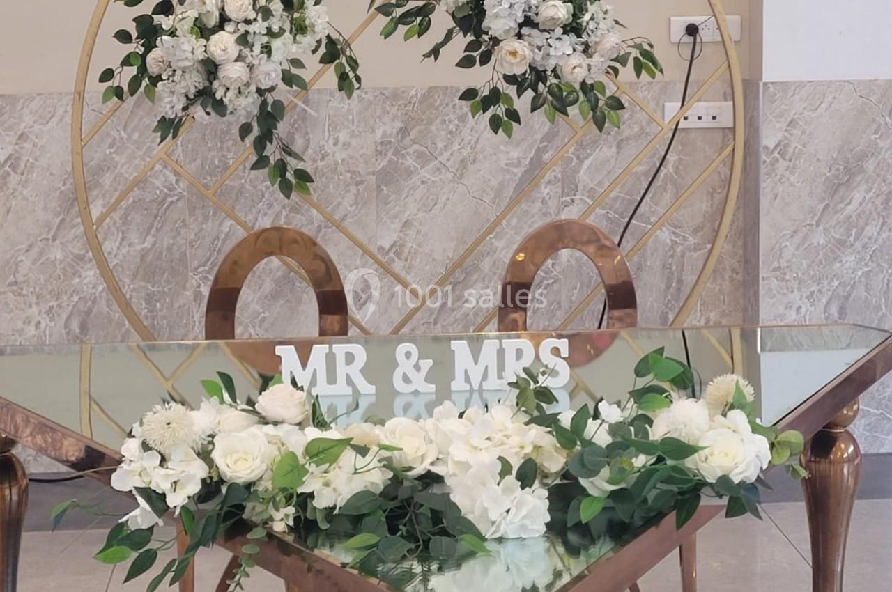 Table décorée pour un mariage avec des fleurs blanches, des feuillages et un panneau ’MR & MRS’ devant un cadre doré.