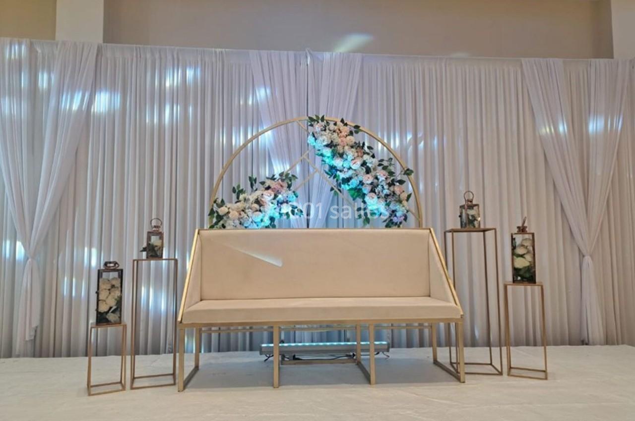 Banc beige devant un rideau blanc plissé, décoré d'un cercle fleuri et de lanternes sur des supports dorés.