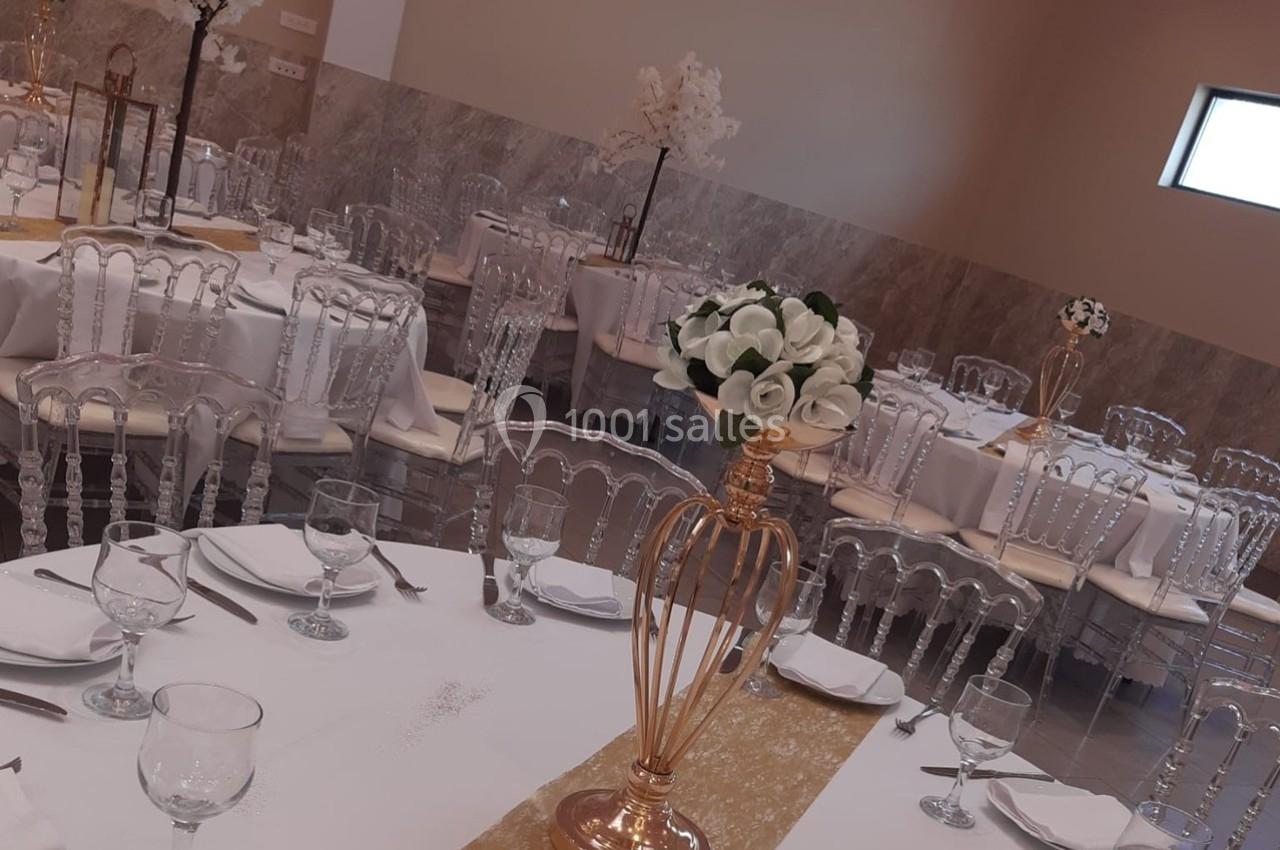 Salle de réception élégamment décorée avec des tables dressées, des chaises transparentes et des centres de table fleuris.