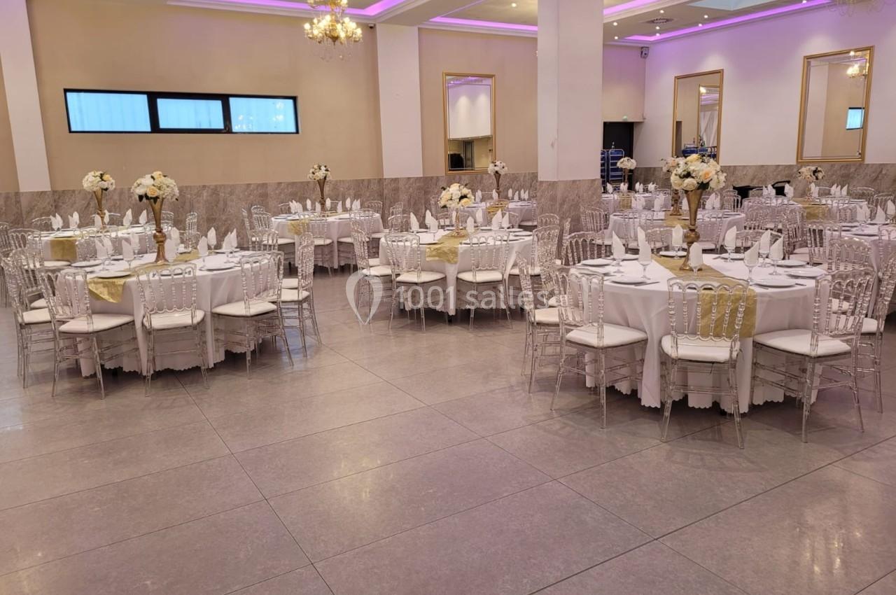 Salle de réception élégante avec tables rondes dressées, chaises transparentes et décorations florales blanches.