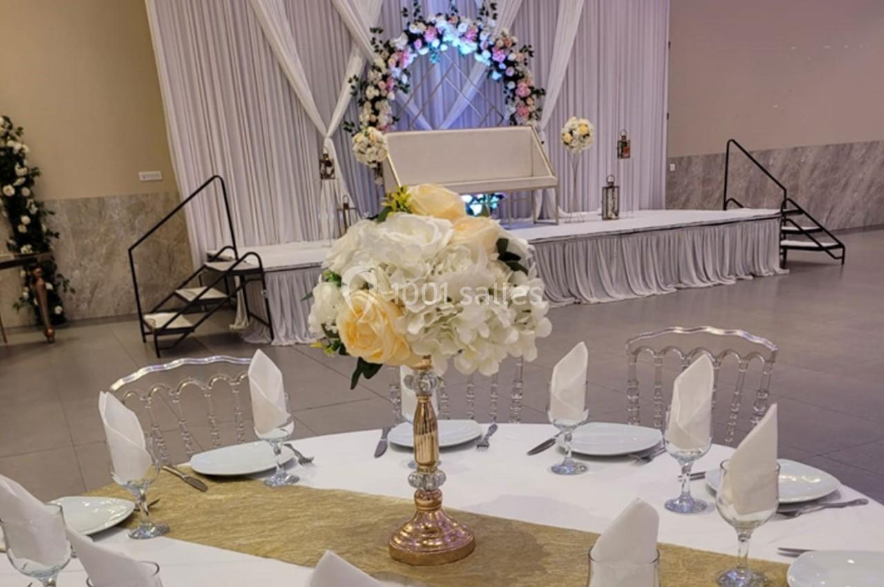Salle de réception décorée avec une table dressée, un centre de table floral et une arche fleurie en arrière-plan.