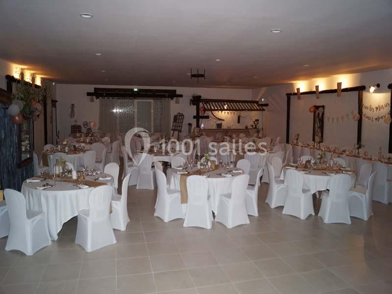 Salle de réception décorée avec des tables rondes, nappes blanches et chaises houssées, prête pour un événement.