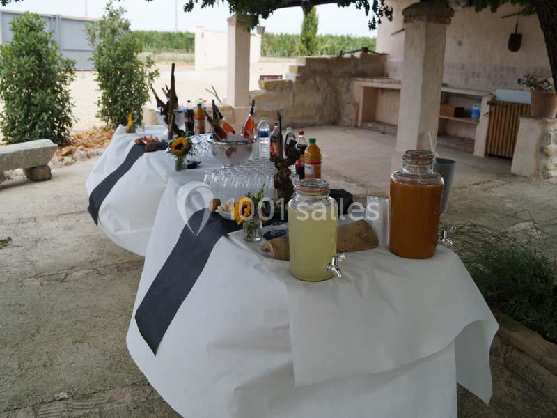 Table dressée en extérieur avec boissons, verres et décorations florales, sous un abri près d'un jardin.