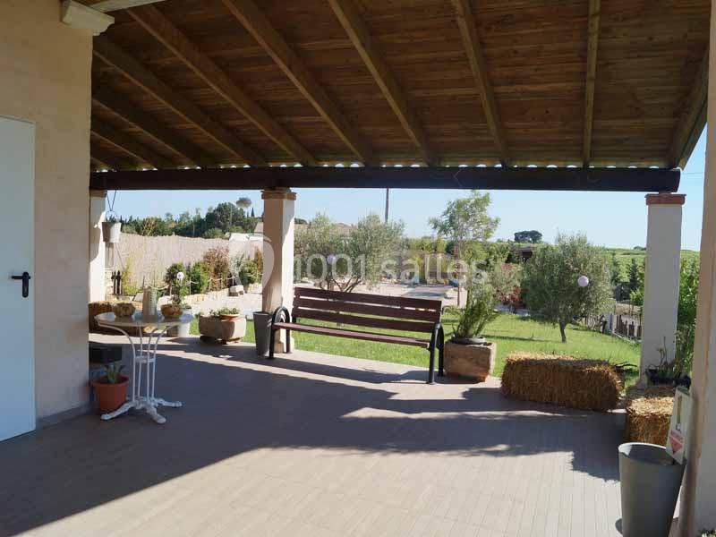Terrasse couverte avec banc en bois, vue sur un jardin arboré et des champs sous un ciel dégagé.