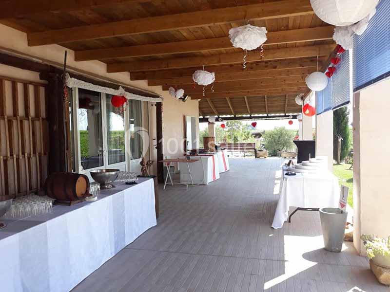 Terrasse couverte aménagée pour un événement, avec tables dressées, décorations suspendues et vue sur un jardin.