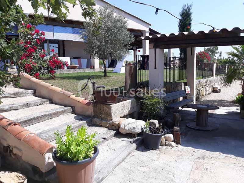Cour extérieure avec terrasse ombragée, plantes en pot, escaliers en pierre et vue sur un jardin fleuri.