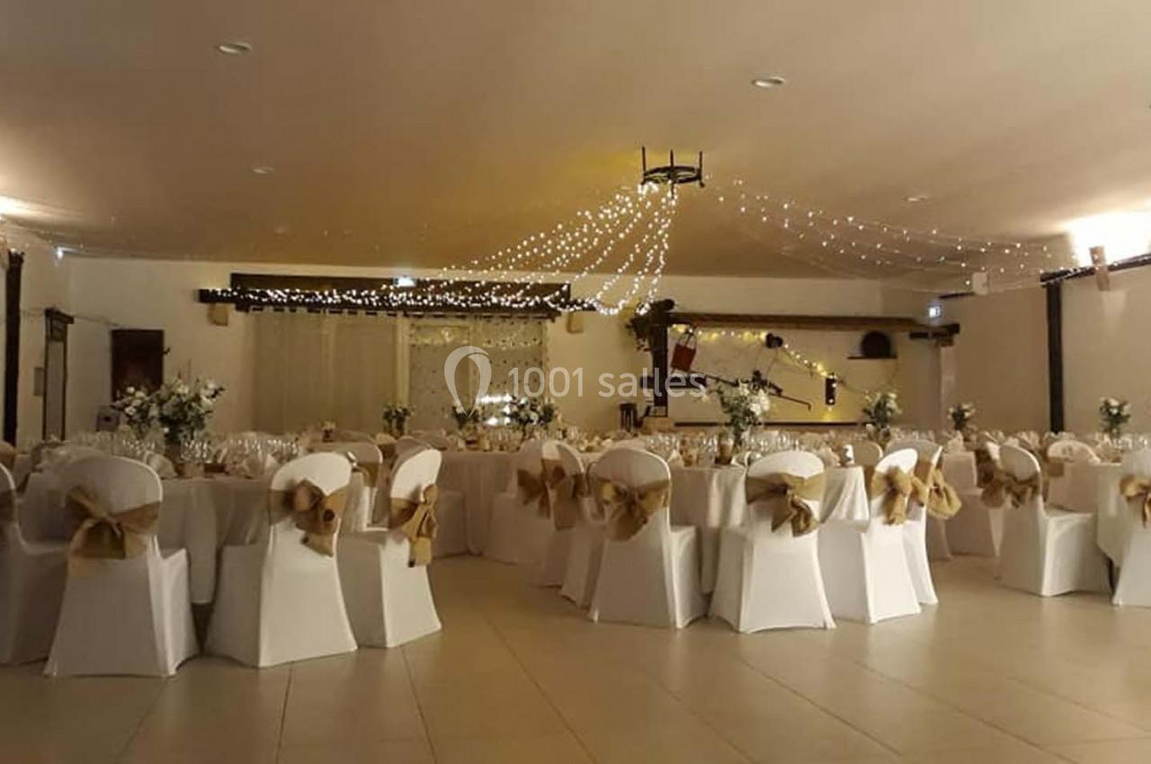 Salle de réception décorée avec des chaises blanches ornées de nœuds dorés et des guirlandes lumineuses suspendues.
