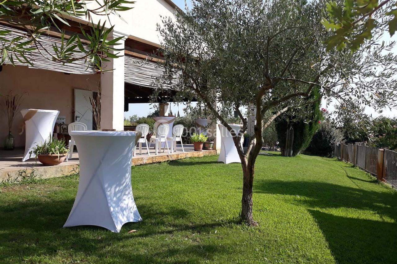 Jardin avec pelouse, tables hautes couvertes de nappes blanches et terrasse ombragée en arrière-plan.