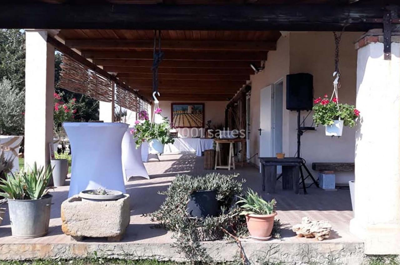 Terrasse couverte avec plantes en pots, table haute blanche et décoration suspendue, dans un cadre extérieur ensoleillé.