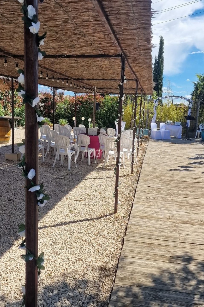 Pergola décorée de fleurs blanches ombrageant des tables et chaises en plastique sur un sol en gravier, avec allée en bois.