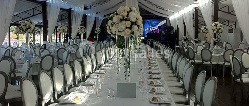 Salle de réception décorée avec des tables alignées, nappes blanches, chaises élégantes et arrangements floraux.