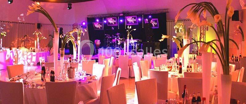 Salle de réception décorée avec des tables rondes, des chaises blanches et des arrangements floraux, éclairée par des…