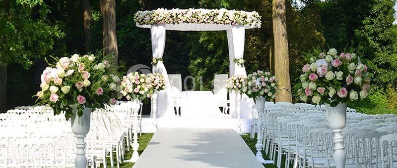 Allée centrale décorée de fleurs blanches et roses menant à une arche de mariage dans un jardin verdoyant.
