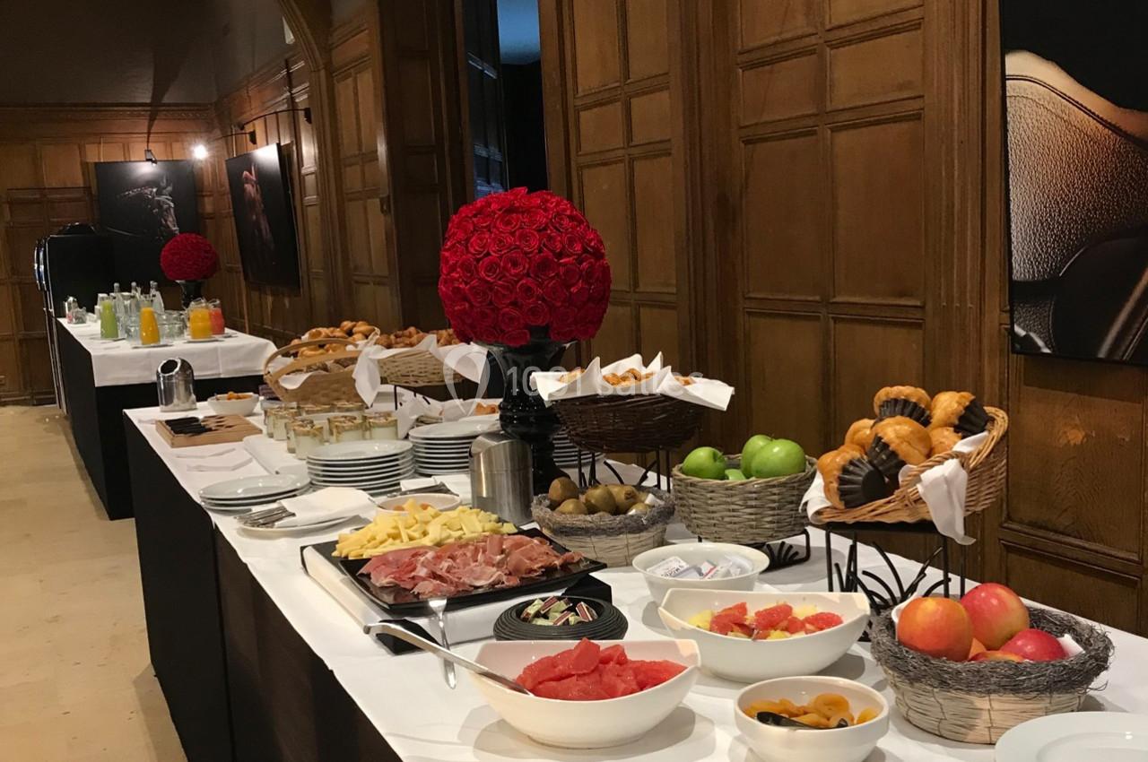 Buffet avec viennoiseries, fruits frais, charcuterie, fromage et boissons disposés sur une table dans une salle en bois.
