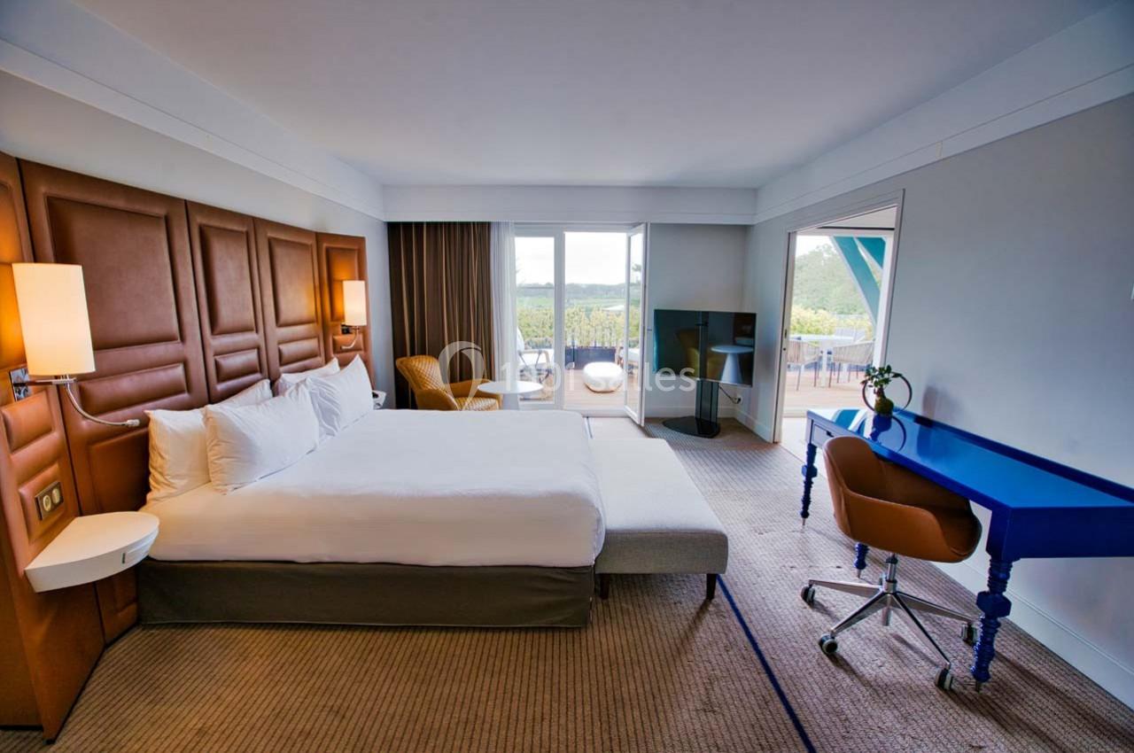 Chambre d'hôtel lumineuse avec lit double, bureau bleu, fauteuil, télévision et balcon offrant une vue sur l'extérieur.