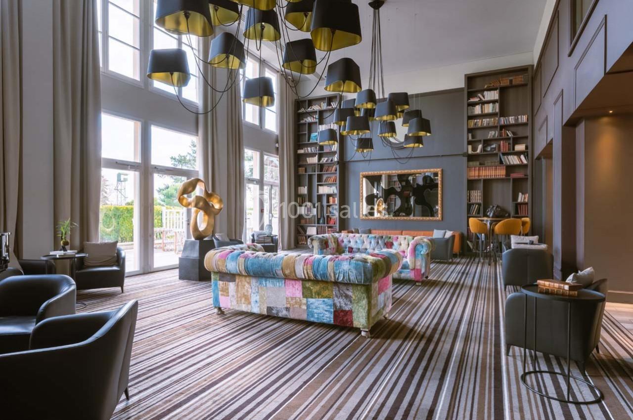 Salon moderne avec canapé coloré, fauteuils noirs, grande bibliothèque, lustres suspendus et larges baies vitrées.