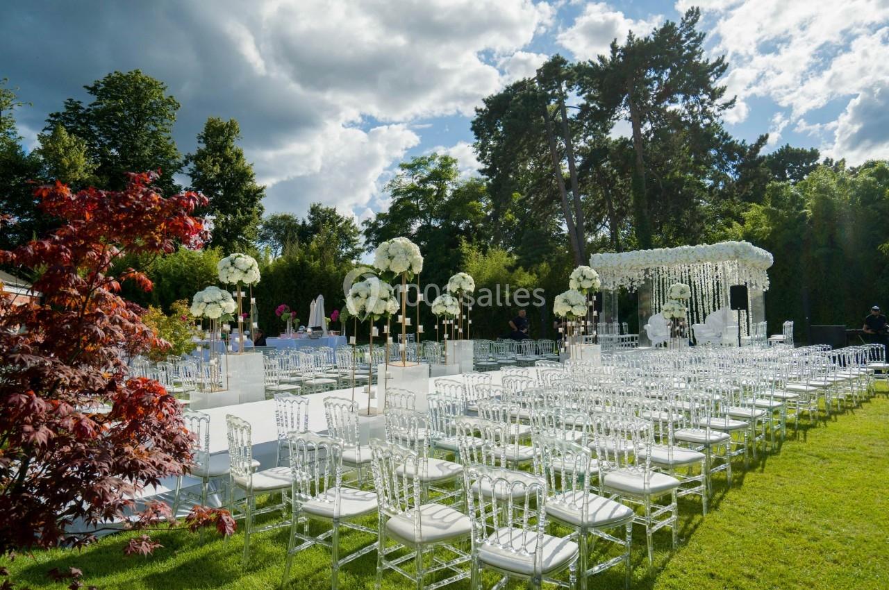 Chaises transparentes disposées en rangées sur une pelouse, avec des décorations florales blanches et une arche en arrière…
