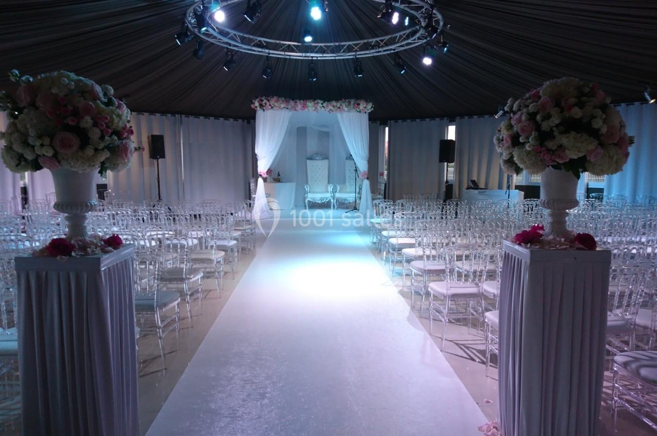 Allée centrale décorée pour une cérémonie, entourée de chaises transparentes et ornée de fleurs sous une tente éclairée.