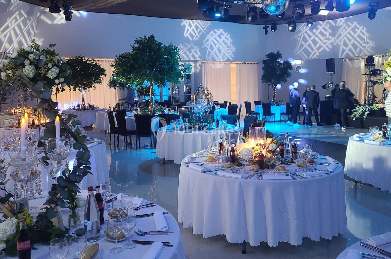 Salle de réception décorée avec des tables rondes dressées, éclairage tamisé et arrangements floraux.