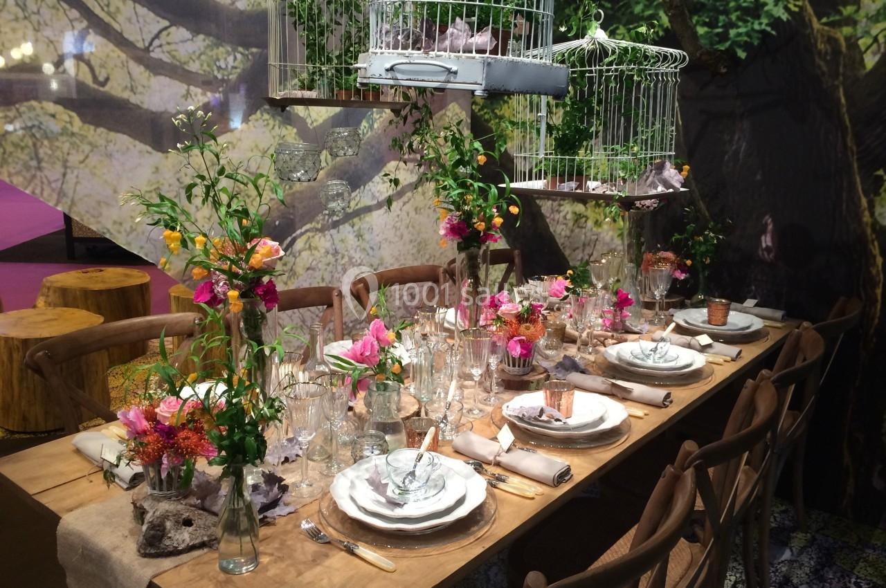 Table en bois décorée avec vaisselle élégante, fleurs colorées et cages à oiseaux suspendues dans un décor naturel.