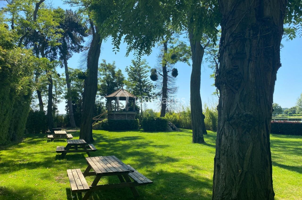 Espace vert avec tables de pique-nique en bois, arbres ombragés et kiosque au fond sous un ciel dégagé.