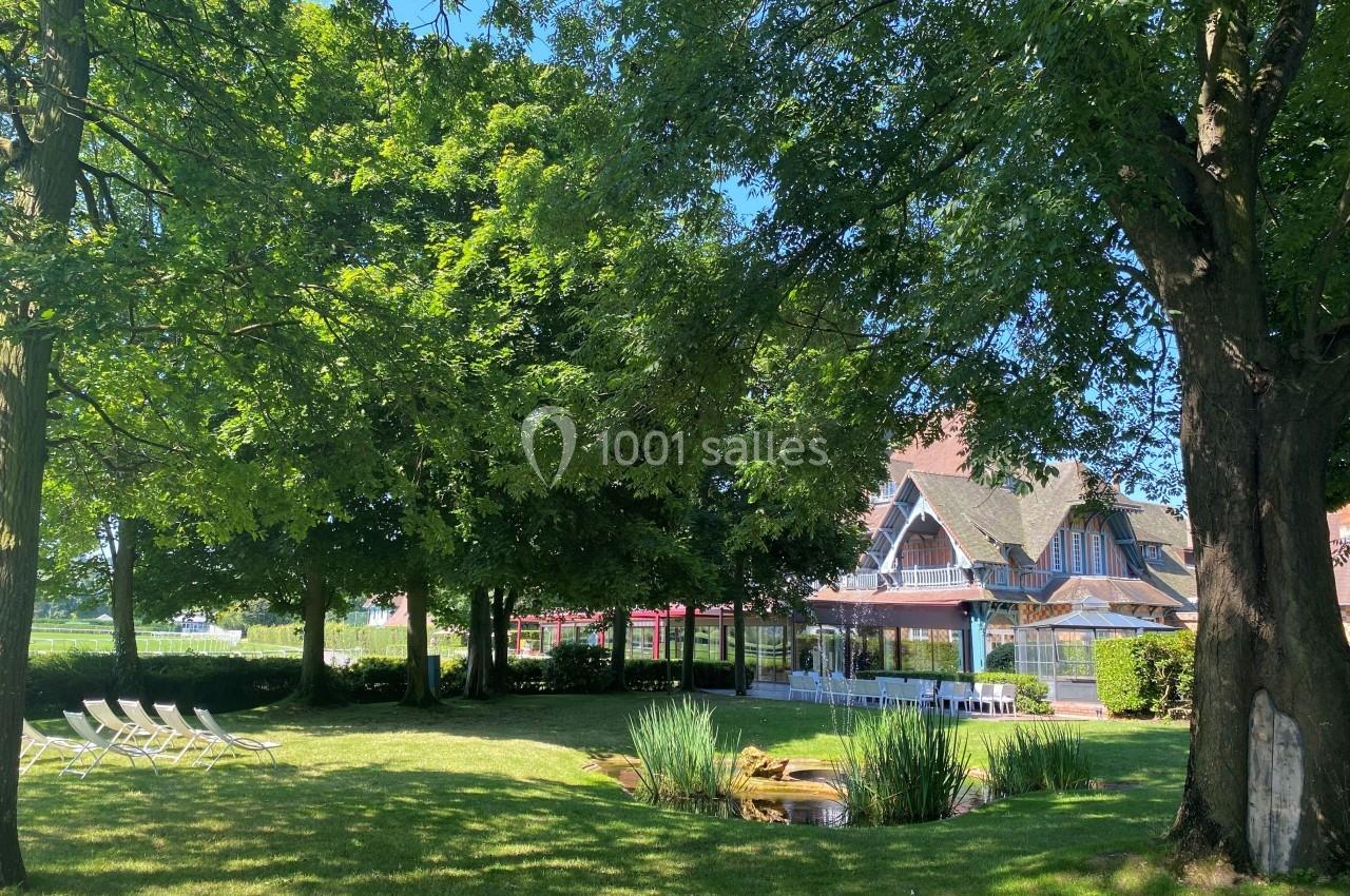 Jardin arboré avec pelouse, chaises longues, petit étang et bâtiment à colombages en arrière-plan.