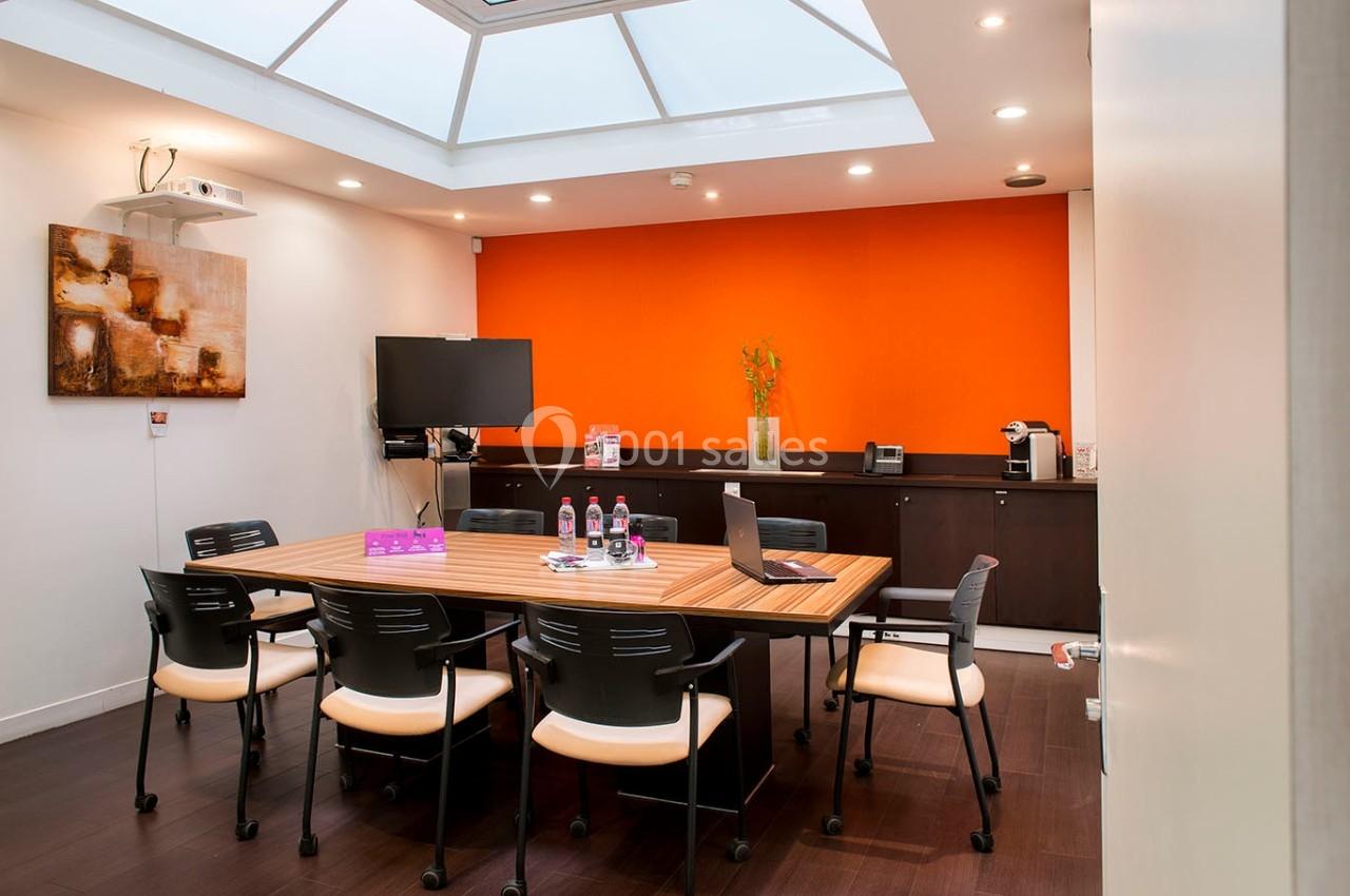 Salle de réunion lumineuse avec table en bois, chaises noires, mur orange, écran, et équipement audiovisuel.