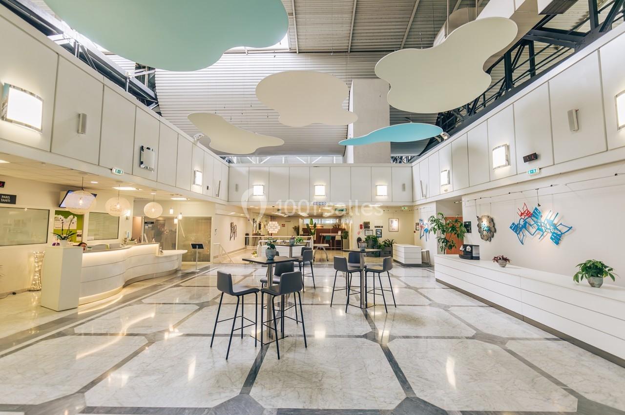 Espace intérieur lumineux avec comptoir d'accueil, tables hautes, plantes et décorations suspendues au plafond.