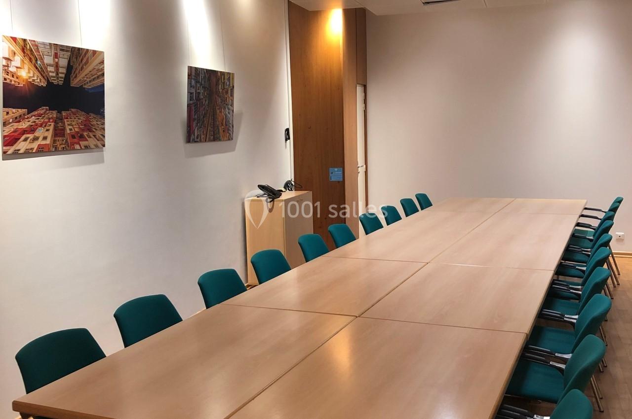 Salle de réunion avec une grande table rectangulaire en bois, entourée de chaises vertes, éclairée par des plafonniers.