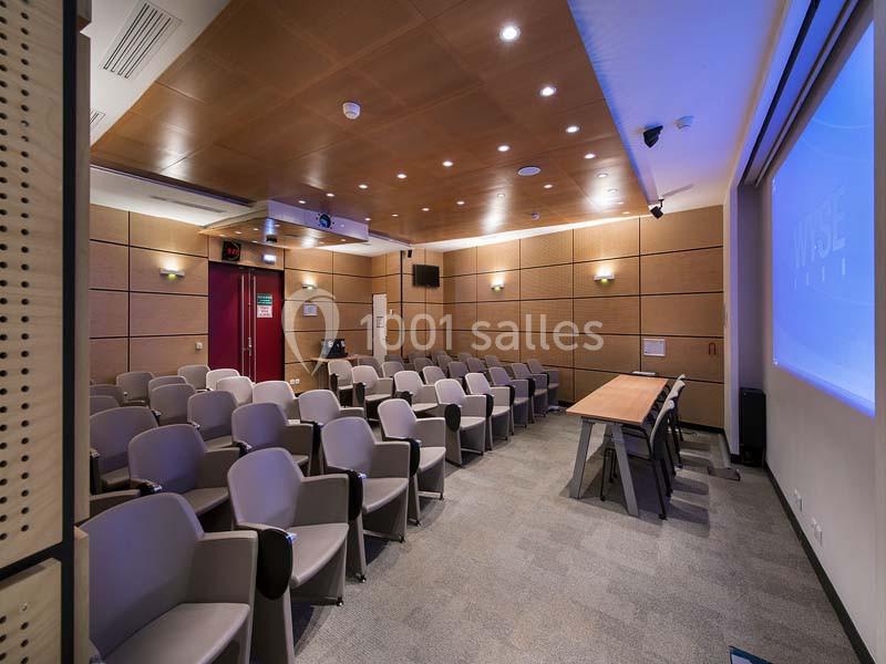 Salle de conférence moderne avec rangées de sièges gris, table en bois à l'avant et écran de projection allumé.