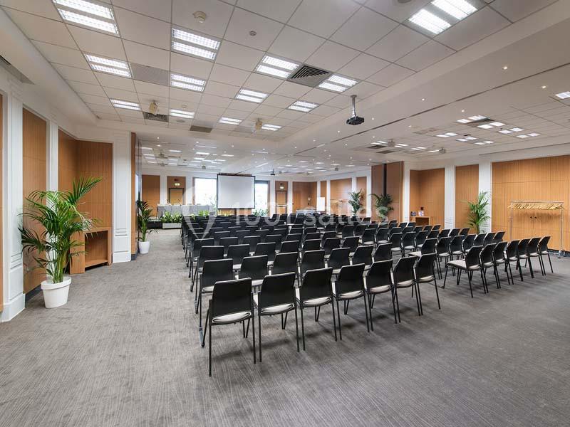 Salle de conférence lumineuse avec rangées de chaises noires, écran de projection et plantes décoratives.