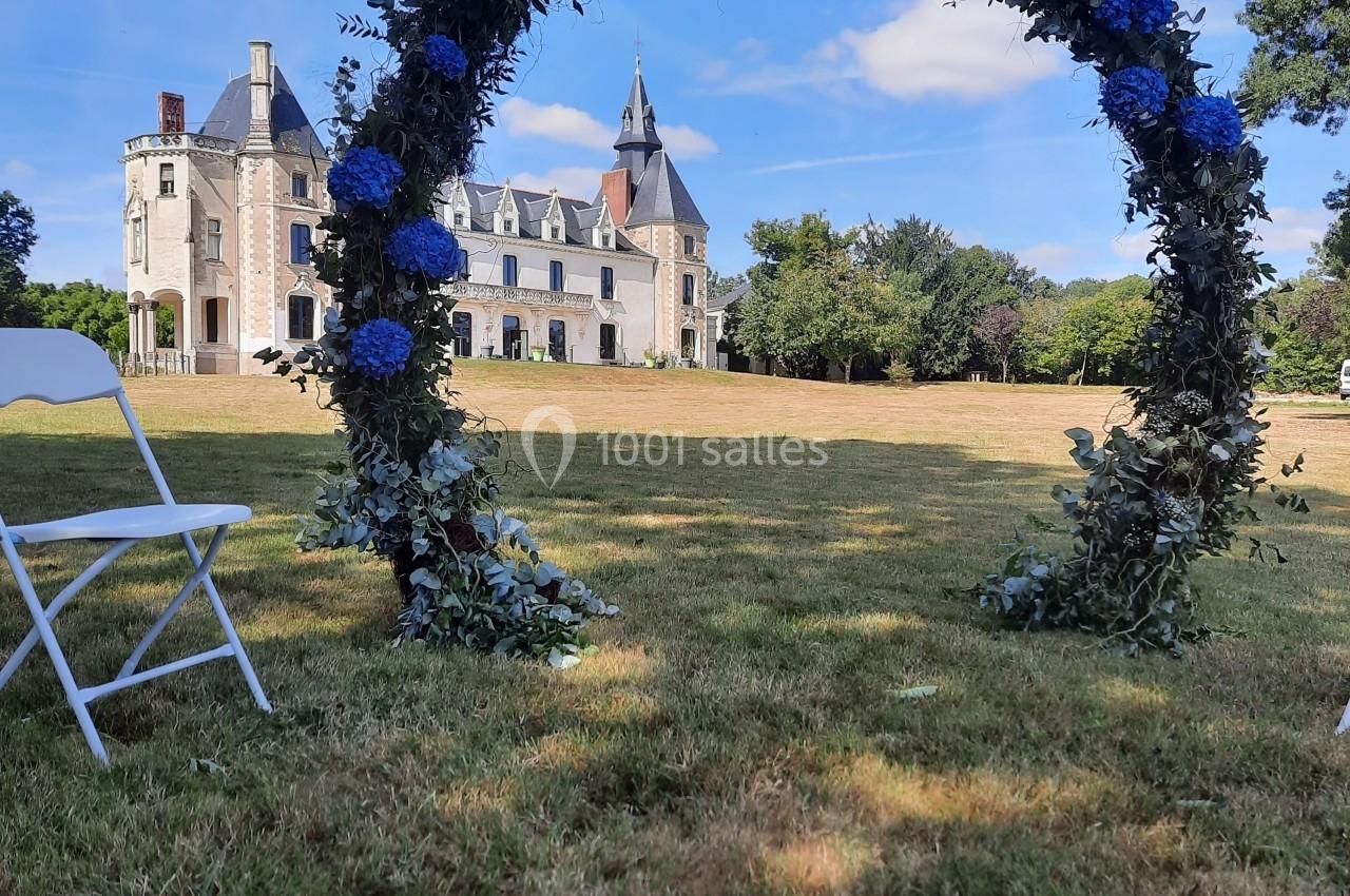 Arc fleuri avec des hortensias bleus sur une pelouse, devant un château sous un ciel bleu.